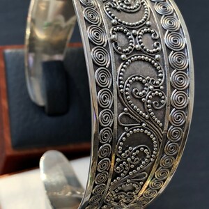 K&ouml;nnte beinhalten: Ein silbernes Armreif-Armband mit einem komplizierten Design aus Spiralen und Punkten. Das Armband ist aus poliertem Silber gefertigt und hat eine glatte, abgerundete Oberfl&auml;che.