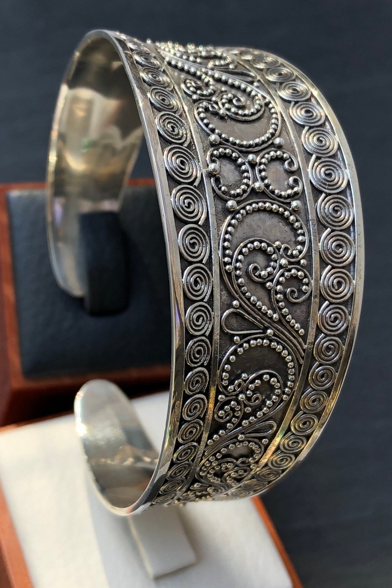 K&ouml;nnte beinhalten: Ein silbernes Armreif-Armband mit einem komplizierten Design aus Spiralen und Punkten. Das Armband ist aus poliertem Silber gefertigt und hat eine glatte, abgerundete Oberfl&auml;che.