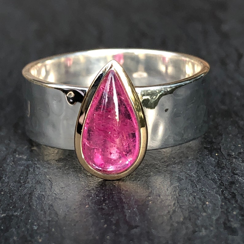 May include: A wide silver ring with a hammered texture. The ring features a teardrop-shaped, vibrant pink gemstone set in a gold bezel. The gemstone is the focal point, showcasing its color and shape.