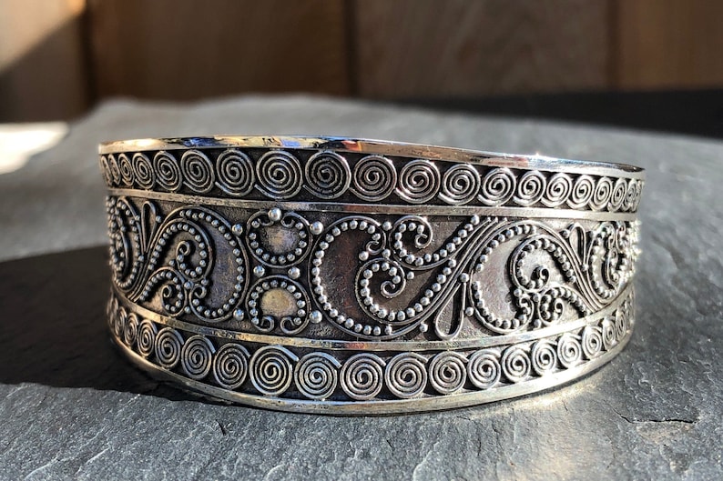 K&ouml;nnte beinhalten: Ein silbernes Armband mit komplizierten, wirbelnden Designs. Das Armband besteht aus einem dicken Metallband und hat eine polierte Oberfl&auml;che.