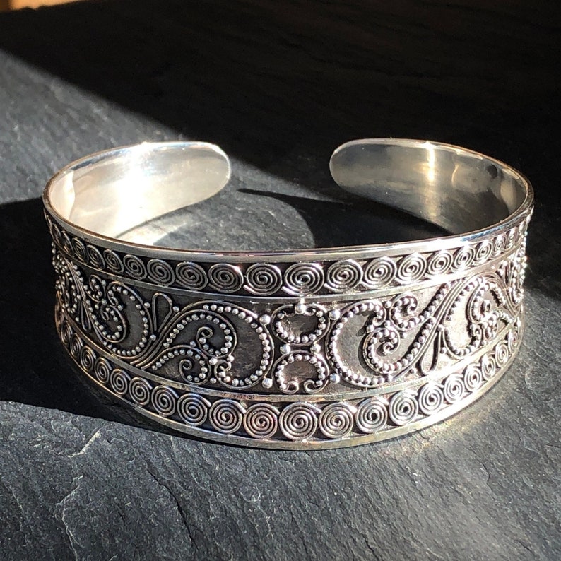 K&ouml;nnte beinhalten: Ein silbernes Armreif-Armband mit einem komplizierten, verzierten Design mit Spiralen und Schn&ouml;rkeln. Das Armband ist offen und verstellbar.
