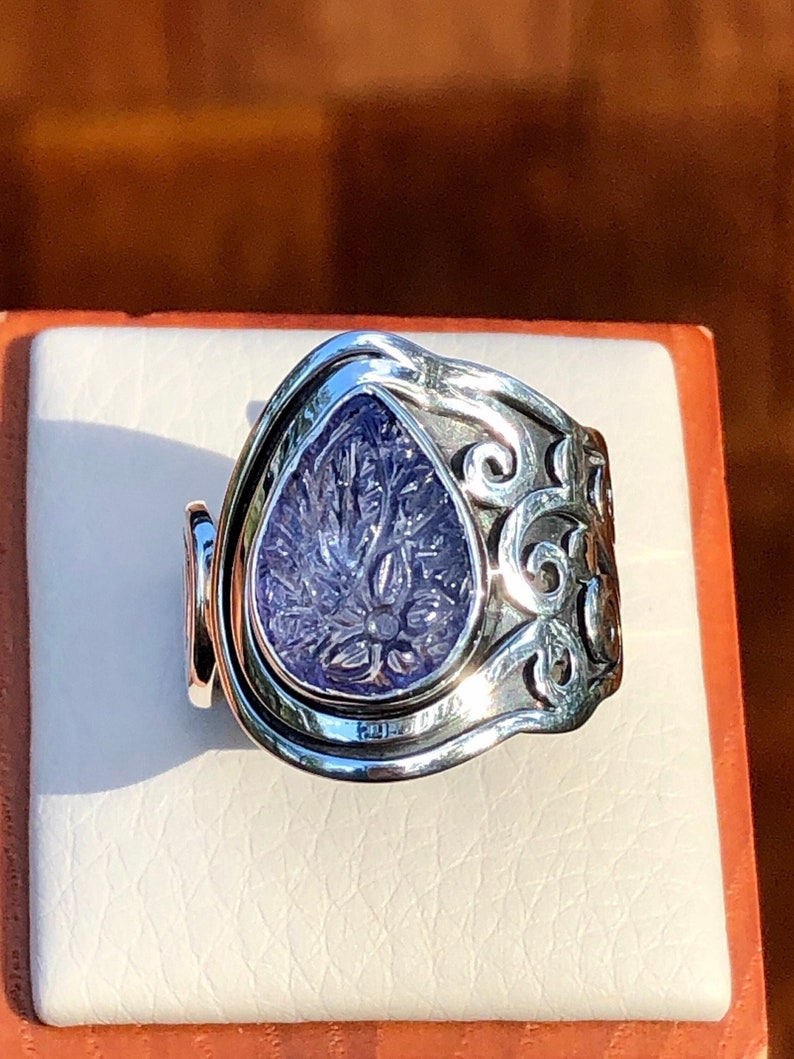 May include: A silver ring with an intricate design featuring a purple gemstone. The gemstone is set in a teardrop shape and has a carved floral pattern.
