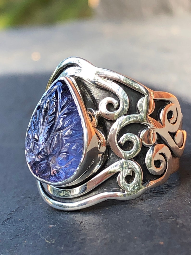 May include: A silver ring with a purple gemstone set in a teardrop shape. The ring has a detailed, ornate design with swirling patterns.