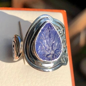 May include: A silver ring with a carved purple gemstone set in a teardrop shape. The ring has a unique design with multiple bands.