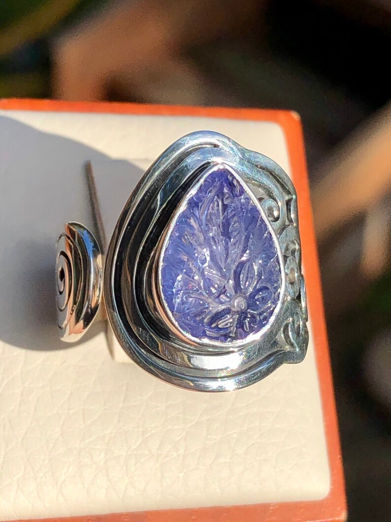 May include: A silver ring with a carved purple gemstone set in a teardrop shape. The ring has a unique design with multiple bands.