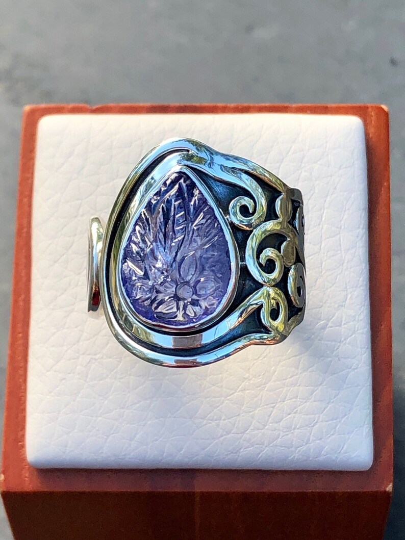 May include: A silver ring with a purple gemstone set in a teardrop shape. The ring has a decorative, scroll-like design around the gemstone.