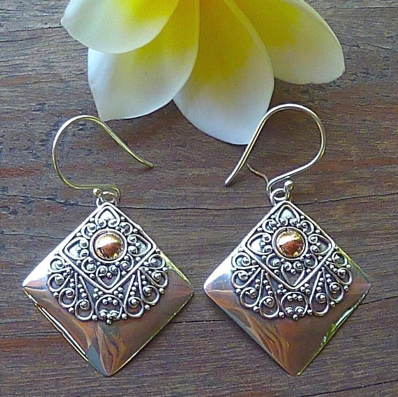 May include: A pair of diamond-shaped earrings with a copper-colored base and ornate silver filigree detailing. Each earring features a gold-colored central accent and a hook closure. The earrings are displayed on a wooden surface.