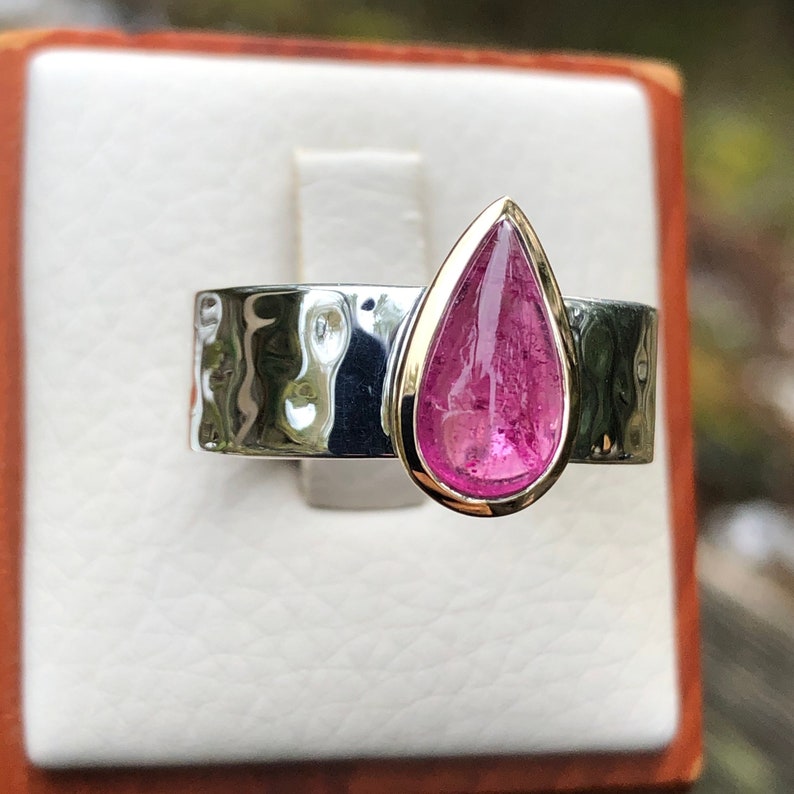 K&ouml;nnte beinhalten: Ein Silberring mit einer breiten, geh&auml;mmerten Ringschiene und einem einzelnen birnenf&ouml;rmigen, durchscheinenden rosa Edelstein in einer goldfarbenen Fassung. Der Stein weist sichtbare Einschl&uuml;sse und eine glatte Oberfl&auml;che auf.