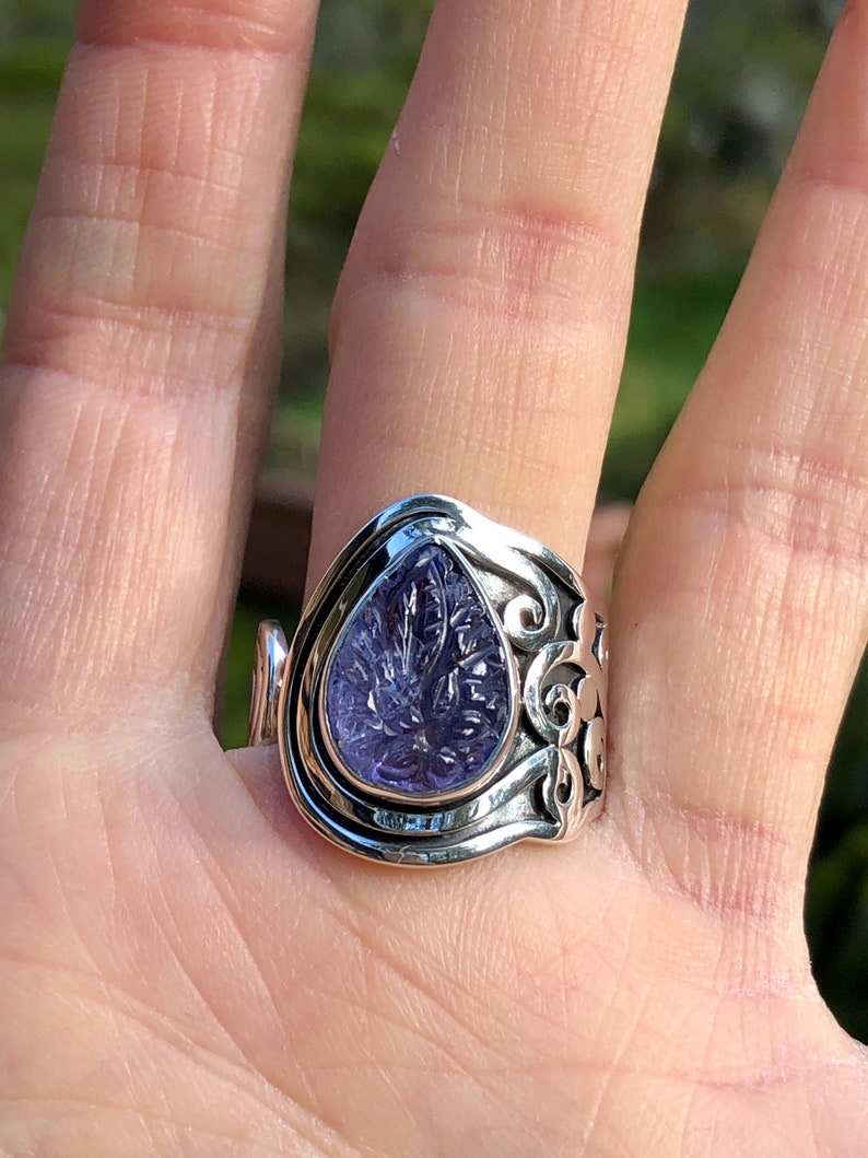 May include: A silver ring with an ornate design features a purple amethyst gemstone in a teardrop shape.