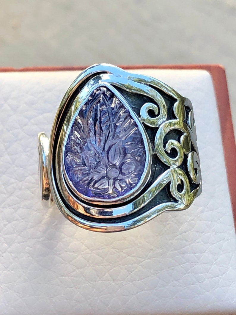 May include: A silver ring with a purple gemstone set in a floral design. The ring is wide and has a unique, curved shape.