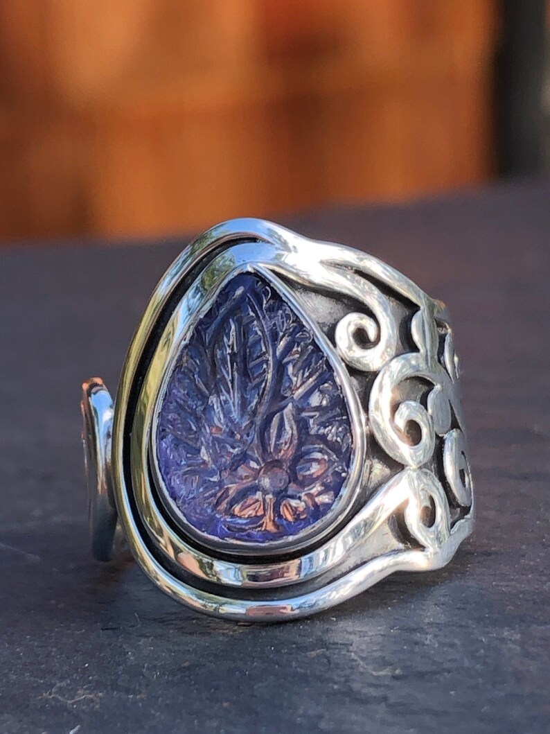 May include: A silver ring with a teardrop-shaped purple stone set in a swirling, ornate design.