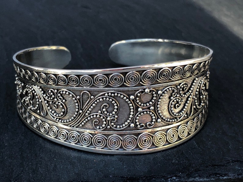 K&ouml;nnte beinhalten: Silberner Armreif mit einem dekorativen Design aus Spiralen und Punkten. Das Armband ist offen und verstellbar.