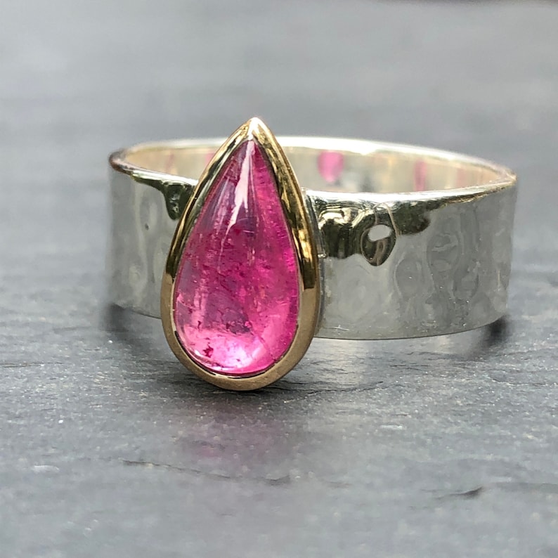 May include: A silver ring with a hammered texture and a teardrop-shaped, vibrant pink gemstone set in a gold bezel. The ring's band is wide and has a textured, hammered finish, creating a contrast with the smooth gemstone.