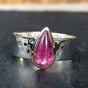 K&ouml;nnte beinhalten: Ein silberner Ring mit geh&auml;mmertem Muster und einer Goldfassung. Der Ring hat einen gro&szlig;en, tropfenf&ouml;rmigen, rosa Edelstein. Der Edelstein ist der Mittelpunkt, gesetzt gegen das kontrastierende Silberband.