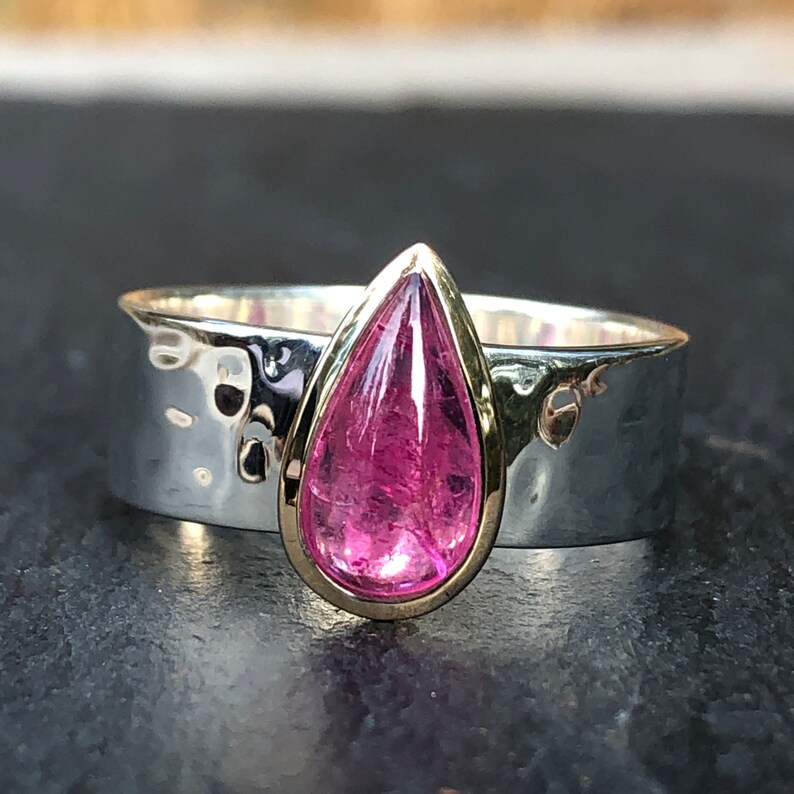 K&ouml;nnte beinhalten: Ein silberner Ring mit geh&auml;mmertem Muster und einer Goldfassung. Der Ring hat einen gro&szlig;en, tropfenf&ouml;rmigen, rosa Edelstein. Der Edelstein ist der Mittelpunkt, gesetzt gegen das kontrastierende Silberband.