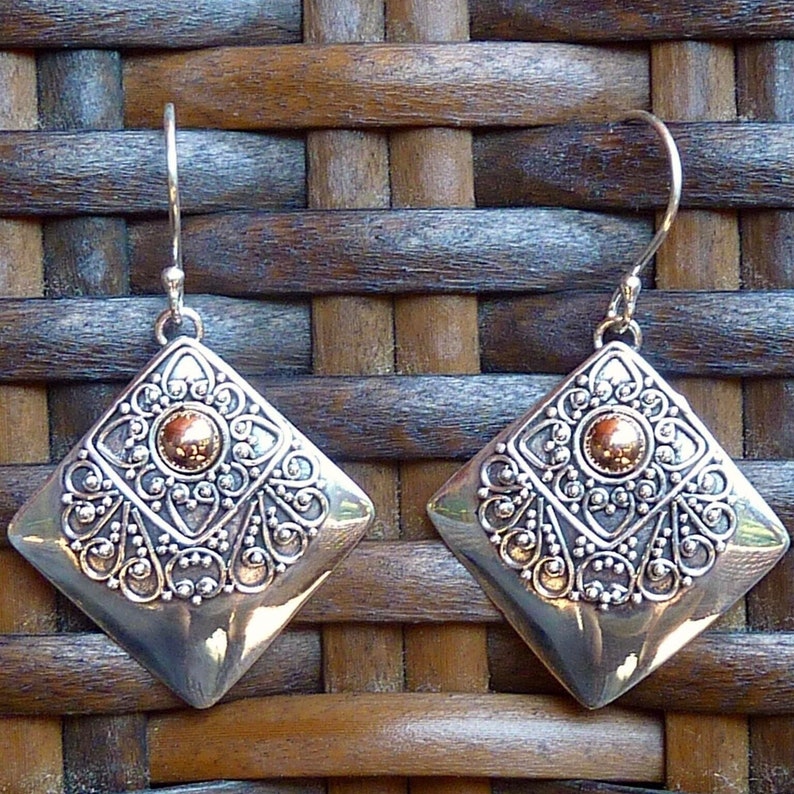 May include: A pair of silver-toned, diamond-shaped earrings with intricate filigree detailing. Each earring features a central, circular, gold-colored accent. The earrings have a hook closure and are displayed against a woven background.