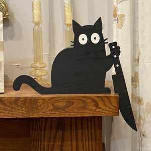 May include: A black wooden cat silhouette with large white eyes, holding a large knife. The cat is sitting on a wooden surface. The cat's tail is curled. The background includes candles and a floral curtain.