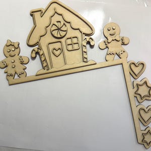 Pode incluir: Decoração de canto em madeira com uma casa de pão de mel e bonecos de pão de mel. A casa tem uma janela em forma de coração e detalhes de bengalas de doces. Formas adicionais incluem corações e estrelas.