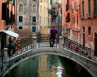 May include: A scenic view of a canal in Venice, Italy, with colorful buildings lining the water. A person stands on a stone bridge with ornate iron railings, looking out over the water. The buildings are in shades of orange, yellow, and gray.