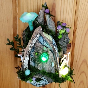Puede incluir: Una casa de hadas en miniatura hecha de madera y musgo con una luz verde brillante en el interior. La casa tiene una pequeña puerta y un techo en forma de hongo. Hay pequeños hongos y hojas verdes alrededor de la casa.