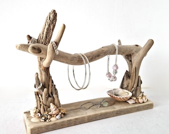 May include: A driftwood jewelry display stand featuring a horizontal branch supported by two vertical pieces on a rectangular wooden base. The base and supports are decorated with small seashells. It holds two silver hoop bracelets, a beaded bracelet, and rings.