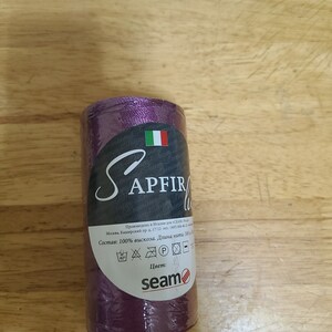 Puede incluir: Una bobina de hilo morado con la etiqueta "SAPFIRE" y el texto "Состав: 100% вискоза. Длина нитки: 500 м" y el logotipo "seam".