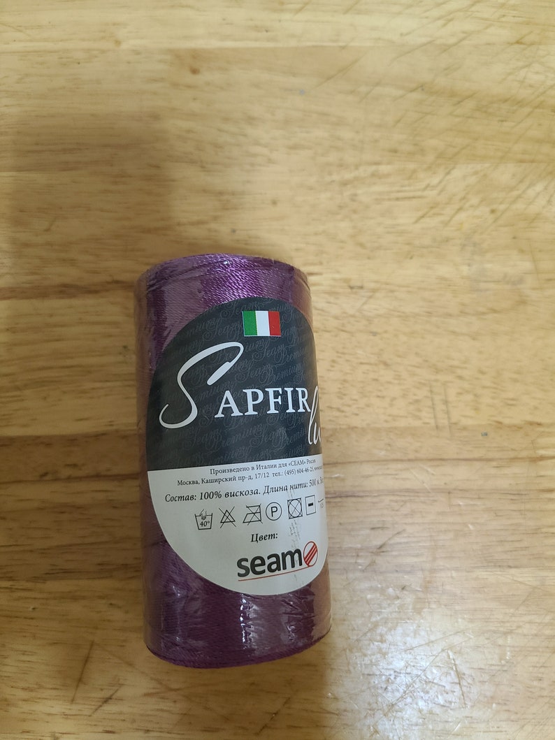 Puede incluir: Una bobina de hilo morado con la etiqueta "SAPFIRE" y el texto "Состав: 100% вискоза. Длина нитки: 500 м" y el logotipo "seam".