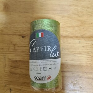 Puede incluir: Una bobina de hilo verde etiquetada "APFIR lux" con el texto "100% viscosa. Longitud: 500 m. Peso: 100 g." e instrucciones de cuidado. La bobina est&aacute; envuelta en pl&aacute;stico transparente.