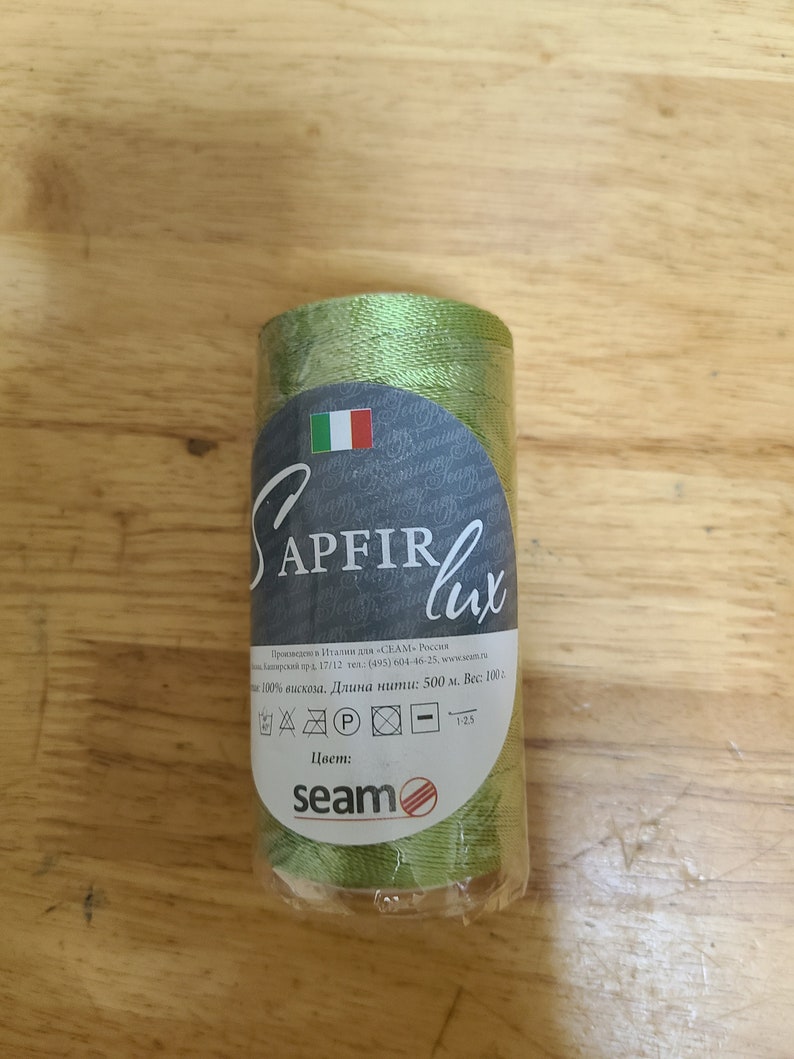 Puede incluir: Una bobina de hilo verde etiquetada "APFIR lux" con el texto "100% viscosa. Longitud: 500 m. Peso: 100 g." e instrucciones de cuidado. La bobina est&aacute; envuelta en pl&aacute;stico transparente.