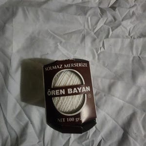 Pode incluir: Uma embalagem castanha e branca de fio mercerizado da marca Ören Bayan. A embalagem tem um design circular com o nome da marca e o texto "NET 100 gramas". O fio é branco e parece estar bem enrolado.