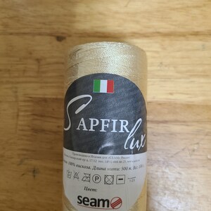 Puede incluir: Una bobina de hilo beige con la etiqueta "SAPFIR lux" y el texto "100% viscosa. Longitud del hilo: 500 m. Peso: 100 g" y "seam" con un logotipo de c&iacute;rculo rojo.