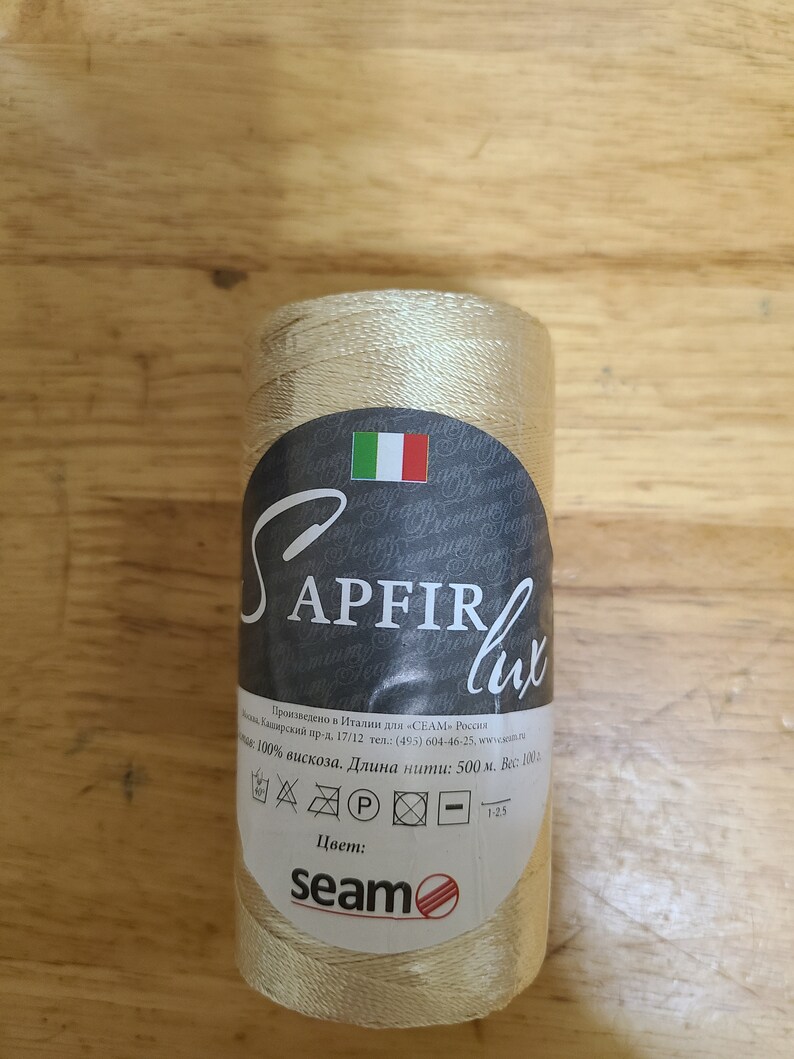 Puede incluir: Una bobina de hilo beige con la etiqueta "SAPFIR lux" y el texto "100% viscosa. Longitud del hilo: 500 m. Peso: 100 g" y "seam" con un logotipo de c&iacute;rculo rojo.