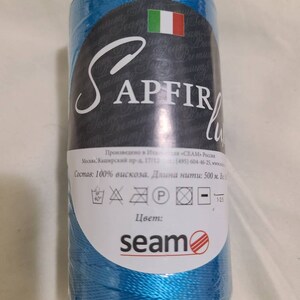 Puede incluir: Una bobina de hilo azul turquesa etiquetada "SAPFIR" con el texto "Состав: 100% вискоза. Длина нити: 500 м. Весь" e instrucciones de cuidado. La bobina est&aacute; envuelta en pl&aacute;stico transparente y tiene un logotipo "seam".