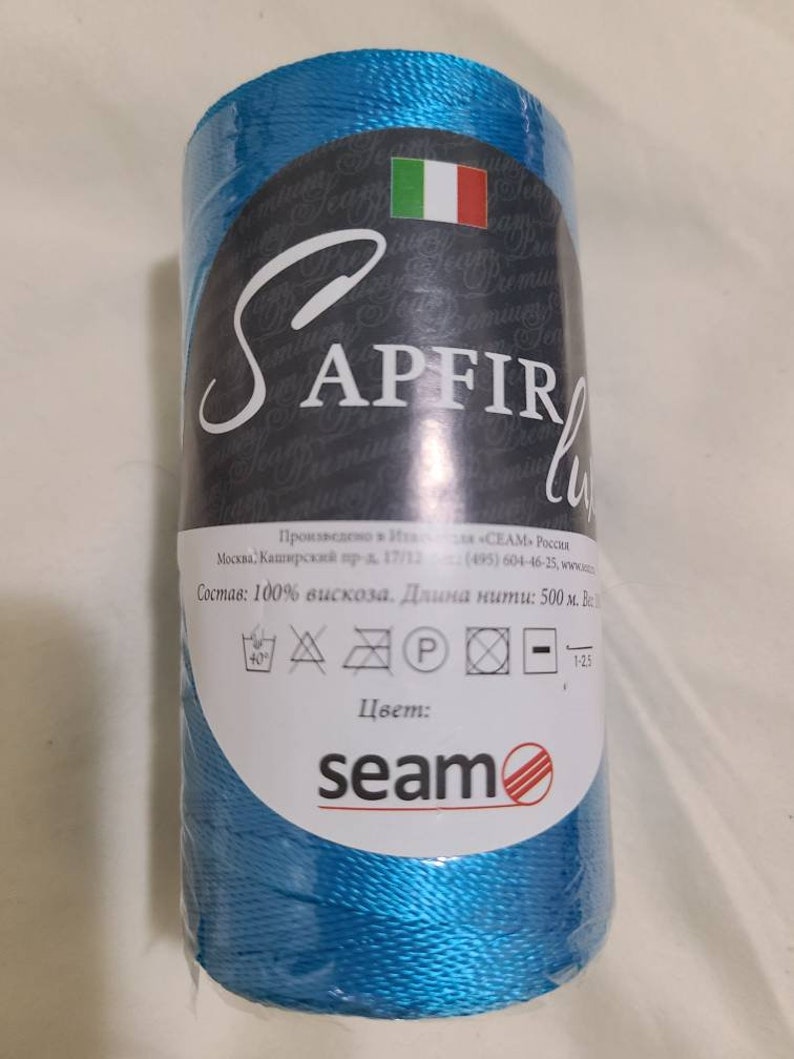 Puede incluir: Una bobina de hilo azul turquesa etiquetada "SAPFIR" con el texto "Состав: 100% вискоза. Длина нити: 500 м. Весь" e instrucciones de cuidado. La bobina est&aacute; envuelta en pl&aacute;stico transparente y tiene un logotipo "seam".