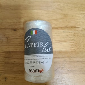 Puede incluir: Una bobina de hilo blanco etiquetada "Sapfir lux" con una peque&ntilde;a bandera italiana e instrucciones de cuidado. La bobina est&aacute; hecha de 100% viscosa y tiene 500 metros de largo. La etiqueta tambi&eacute;n incluye el nombre de la marca "seam" con un logotipo de tres l&iacute;neas rojas.