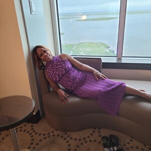 Puede incluir: Una mujer con un vestido morado est&aacute; sentada en una chaise longue marr&oacute;n en una habitaci&oacute;n de hotel. La chaise longue est&aacute; situada frente a una gran ventana con vistas a una masa de agua y una zona verde. Los pies de la mujer descansan en la chaise longue y sus manos descansan en la chaise longue. La mujer est&aacute; mirando a la c&aacute;mara.