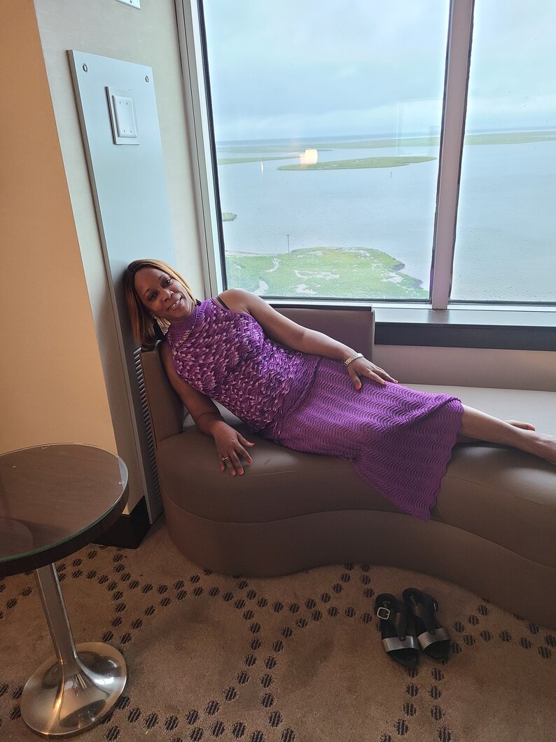 Puede incluir: Una mujer con un vestido morado est&aacute; sentada en una chaise longue marr&oacute;n en una habitaci&oacute;n de hotel. La chaise longue est&aacute; situada frente a una gran ventana con vistas a una masa de agua y una zona verde. Los pies de la mujer descansan en la chaise longue y sus manos descansan en la chaise longue. La mujer est&aacute; mirando a la c&aacute;mara.