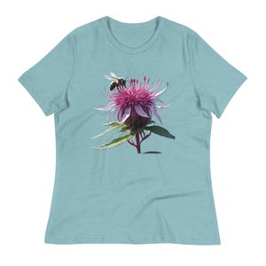 Może przedstawiać: Jasnoniebieska koszulka z wizerunkiem pszczoły na różowym kwiatku ostu. Kwiat ma delikatne płatki i zielone liście. Pszczoła jest w locie, dodając dynamiki do projektu.