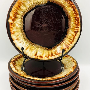 May include: A stack of five round ceramic plates featuring a dark brown center and a wide, textured rim with a drip-glaze pattern in shades of cream, tan, and dark brown. The top plate is displayed upright on a black metal wire stand.