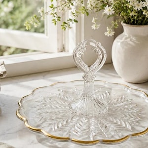 May include: A clear glass serving tray with a scalloped edge and a gold-painted rim. The tray features a central vertical handle with a decorative loop at the top and a pressed leaf-like pattern radiating from the centre base of the handle.