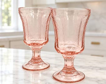 May include: Two matching pale pink glass goblets featuring a textured, embossed floral pattern on the exterior. Each glass has a wide bowl, a short stem, and a circular base, crafted from translucent pink glass with a subtle reflective finish.
