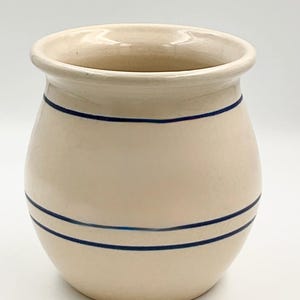 May include: A cream-colored ceramic crock with a rounded body and a wide, flared rim. The exterior features two distinct horizontal navy blue stripes, with one single stripe near the top and a pair of parallel stripes encircling the lower section.