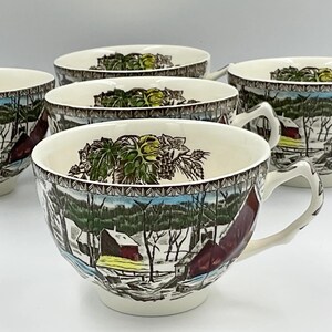 May include: A set of four cream-colored ceramic teacups featuring a winter landscape scene with red barns, bare trees, and snow-covered ground. The interior rim of each cup is decorated with a detailed green leaf and pinecone pattern.