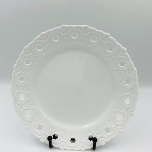 May include: A white ceramic plate with a heart-shaped design around the rim. The plate has a smooth, glossy surface and is displayed on a black stand. The plate's rim features a repeating pattern of open hearts.