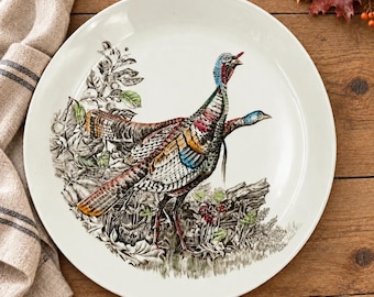 May include: A round white ceramic dinner plate featuring a detailed illustration of two turkeys standing among foliage and branches. The turkeys are rendered in shades of brown, blue, red, and white, with intricate line work throughout the design.