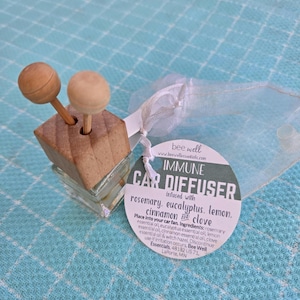 May include: A clear glass car diffuser bottle with a square wooden cap and two wooden ball-topped sticks. A round white tag attached with a white ribbon reads: bee well, IMMUNE CAR DIFFUSER, infused with rosemary, eucalyptus, lemon, cinnamon and clove.