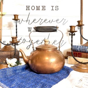 May include: A copper kettle with a wooden handle and lid, flanked by antique candle holders and books. The background features a wooden sign with the text "HOME IS wherever WE are TOGETHER."