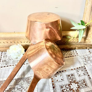 May include: Two antique copper ladles with long wooden handles. The ladles are nested together and have a worn, aged patina.