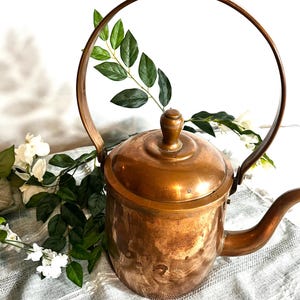 May include: A vintage copper teapot with a large arched handle and a spout. The teapot has a lid with a small knob and is surrounded by green leaves and white flowers. The copper has a warm, aged patina.
