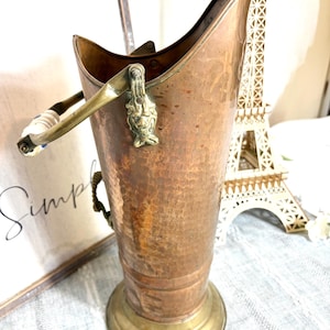 May include: A vintage copper and brass coal scuttle with a hammered texture. The scuttle features a brass handle with a white and blue handle, and a decorative brass accent. The base is a contrasting brass color.
